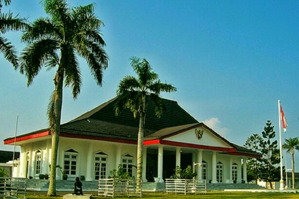 White museum-style building with red accents and columns, set on a grassy lawn with tall palm trees on either side.