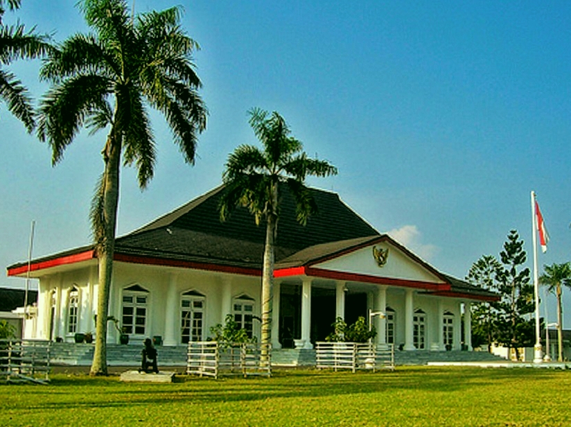 White museum-style building with red accents and columns, set on a grassy lawn with tall palm trees on either side.