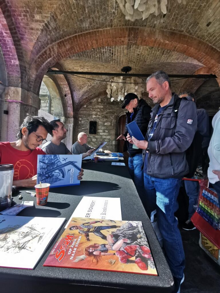 Man at a book signing table signs a comic book as fans stand nearby inside a brick-arched, stone hall; tables display artwork and materials.