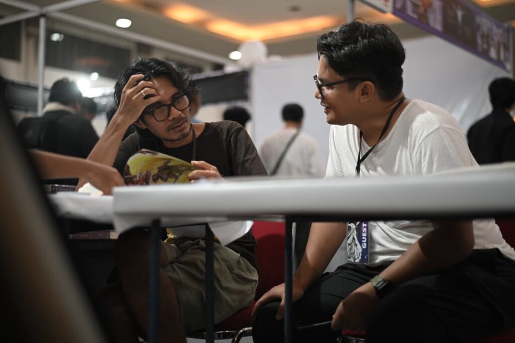 Two men sit at a table at a convention, one studying a magazine with his hand on his head while the other in a white shirt chats nearby.