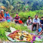 A group of men and boys sit on rocks along a tropical riverbank, watching a colorful ceremonial offering laid out on banana leaves in the foreground, with lush green plants behind them.
