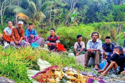 A group of men and boys sit on rocks along a tropical riverbank, watching a colorful ceremonial offering laid out on banana leaves in the foreground, with lush green plants behind them.