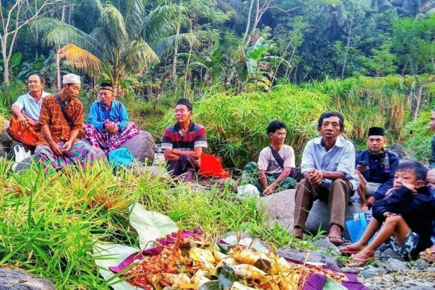 A group of men and boys sit on rocks along a tropical riverbank, watching a colorful ceremonial offering laid out on banana leaves in the foreground, with lush green plants behind them.