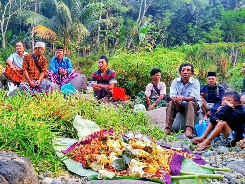 A group of men and boys sit on rocks along a tropical riverbank, watching a colorful ceremonial offering laid out on banana leaves in the foreground, with lush green plants behind them.