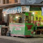 Open-front street shop with colorful banners and a green counter, selling groceries and kitchenware.