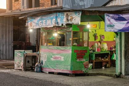 Open-front street shop with colorful banners and a green counter, selling groceries and kitchenware.
