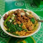 Bowl of stir-fried noodles with leafy greens, meat, and sliced onions on a green patterned table surface.