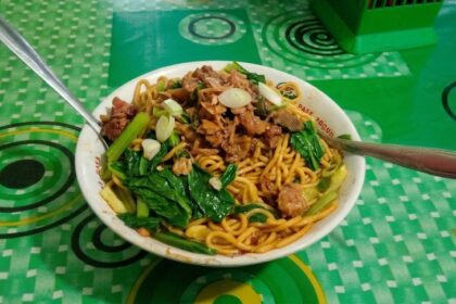 Bowl of stir-fried noodles with leafy greens, meat, and sliced onions on a green patterned table surface.