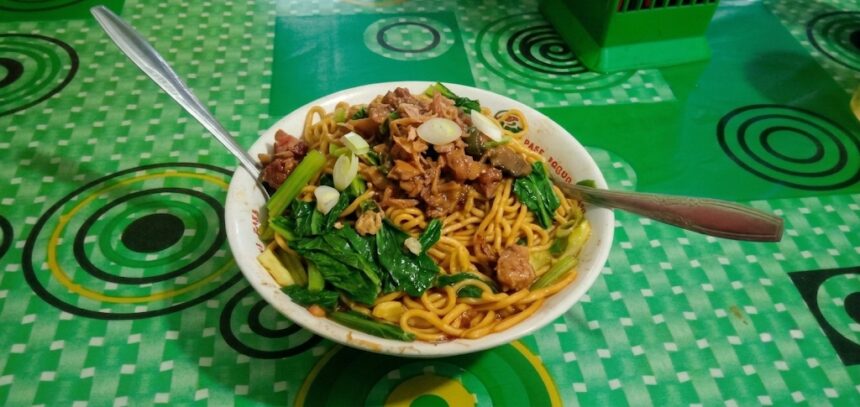 WhatsApp Image 2026-06-11 at 212822 - Mabur.co Bowl of stir-fried noodles with leafy greens, meat, and sliced onions on a green patterned table surface.