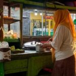 Street food vendor wearing an orange headscarf stirs a pot at a green wooden cart on a street market.