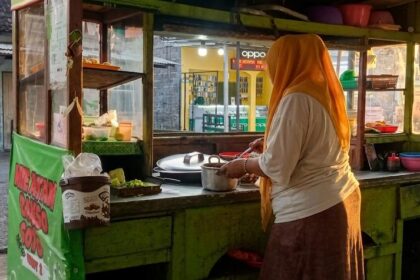 Street food vendor wearing an orange headscarf stirs a pot at a green wooden cart on a street market.