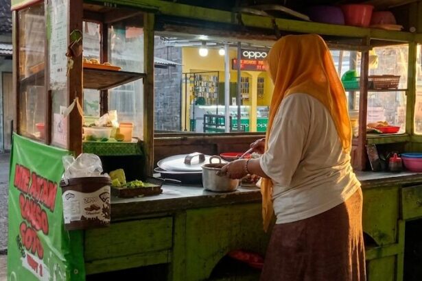 Street food vendor wearing an orange headscarf stirs a pot at a green wooden cart on a street market.