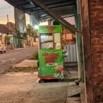 Green street-food cart on a sidewalk advertising mie ayam bakso soto, with a brick wall to the right and a residential street in the background at sunset.
