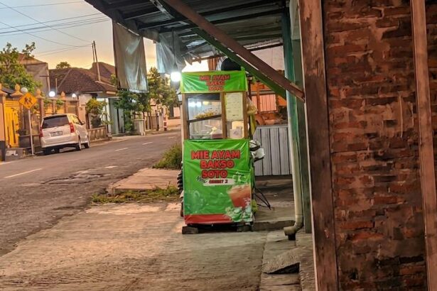 Green street-food cart on a sidewalk advertising mie ayam bakso soto, with a brick wall to the right and a residential street in the background at sunset.