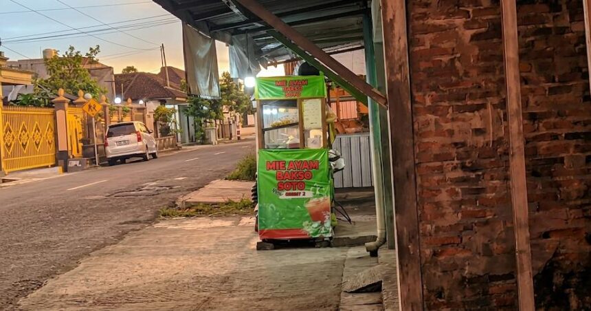 Green street-food cart on a sidewalk advertising mie ayam bakso soto, with a brick wall to the right and a residential street in the background at sunset.