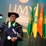 Graduate in a ceremonial gown and cap holding a diploma and a small flag, posing in front of colorful university banners at a graduation ceremony.