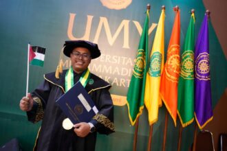 Graduate in a ceremonial gown and cap holding a diploma and a small flag, posing in front of colorful university banners at a graduation ceremony.