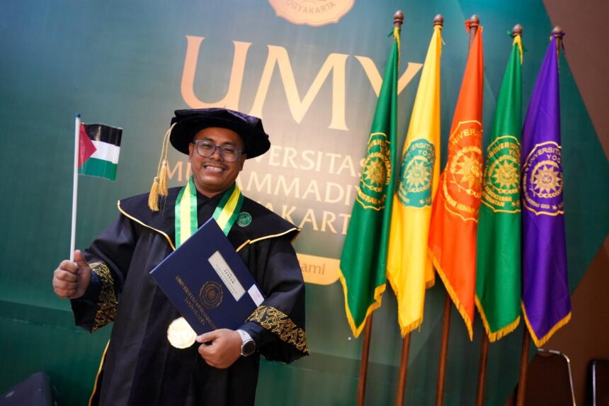 Graduate in a ceremonial gown and cap holding a diploma and a small flag, posing in front of colorful university banners at a graduation ceremony.