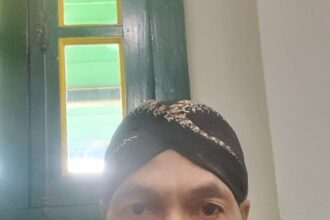 Self-portrait of an older woman wearing a dark patterned head wrap and a blue kurta, indoors near a green-framed window, looking at the camera.
