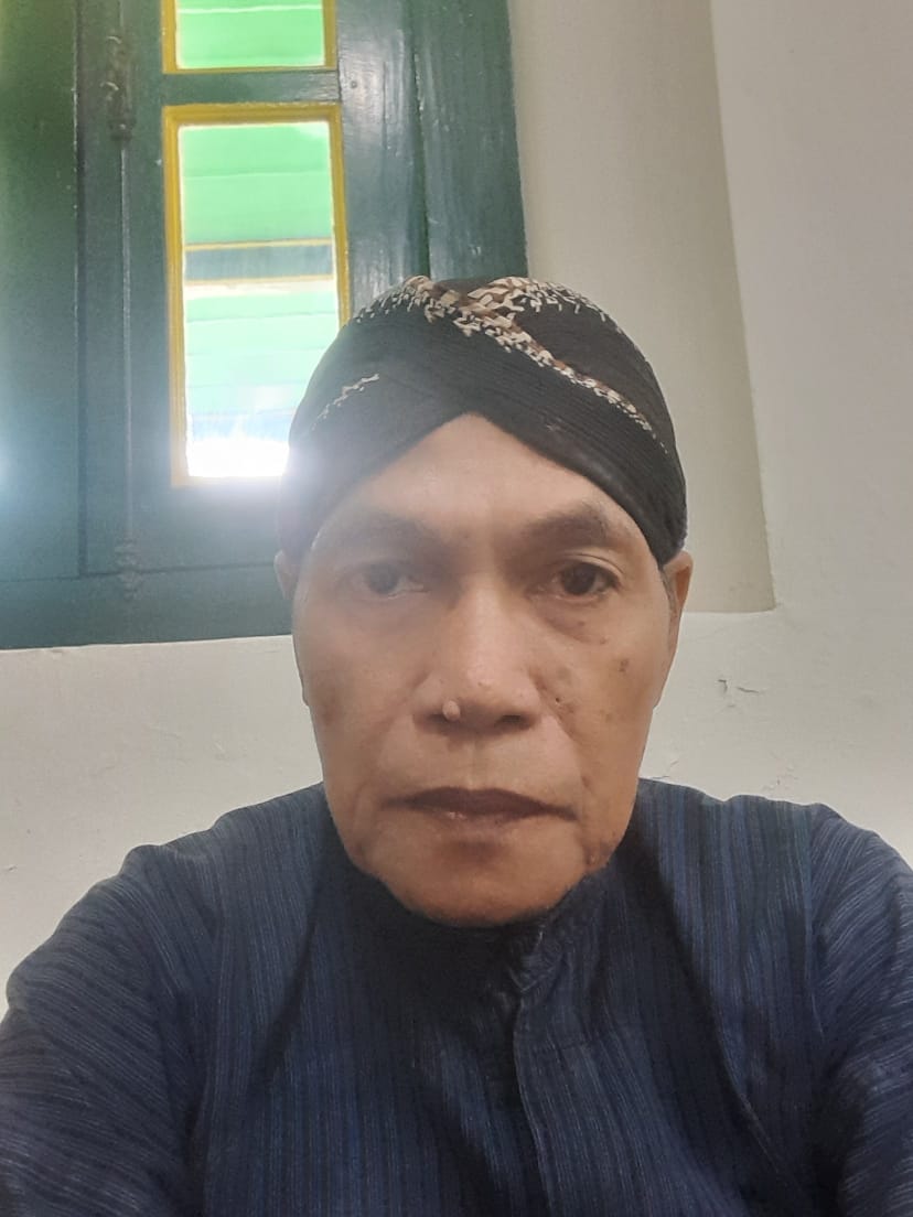 WhatsApp Image 2026-06-15 at 120449 1 - Mabur.co Self-portrait of an older woman wearing a dark patterned head wrap and a blue kurta, indoors near a green-framed window, looking at the camera.