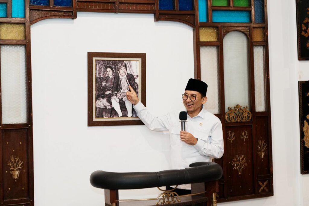 WhatsApp Image 2026-06-20 at 152838 - Mabur.co Speaker in white shirt and black cap presents beside a framed black-and-white photo, pointing at it in a room with decorative wooden panels and stained-glass accents.