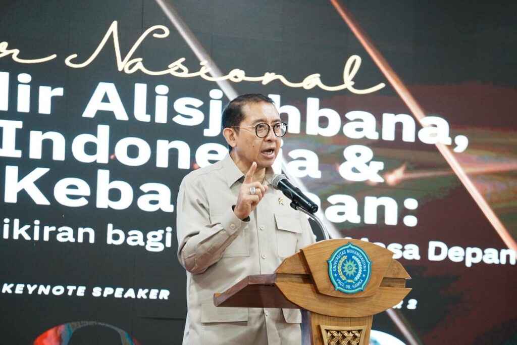 Man in a beige shirt speaks at a wooden podium with a microphone, gesturing with his index finger on stage.