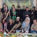 Group of adults posing for a group photo at a cultural ceremony, several holding palm fronds, with ritual food offerings arranged in front.
