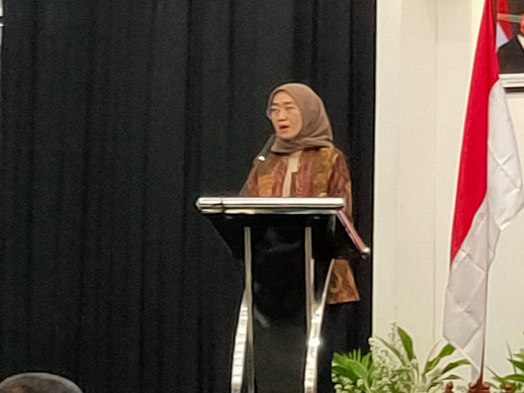 WhatsApp Image 2026-07-08 at 184719 - Mabur.co Woman wearing a hijab speaking at a podium on a stage with a black curtain backdrop and a red-and-white flag nearby.