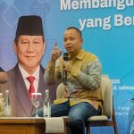 Panelist speaking into a handheld microphone on stage, wearing a batik shirt, with a large portrait and blue banner in the background.