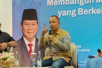 Panelist speaking into a handheld microphone on stage, wearing a batik shirt, with a large portrait and blue banner in the background.