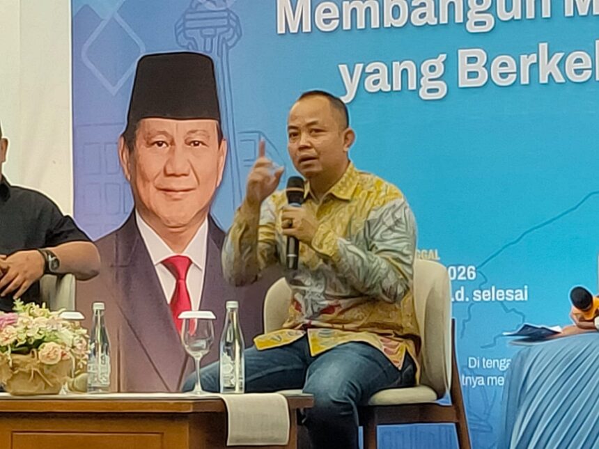 WhatsApp Image 2026-07-08 at 184720 - Mabur.co Panelist speaking into a handheld microphone on stage, wearing a batik shirt, with a large portrait and blue banner in the background.