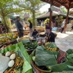 Market scene with banana leaf platters of food, eggs, bananas, and vegetables as people in traditional clothing browse under a wooden pavilion.