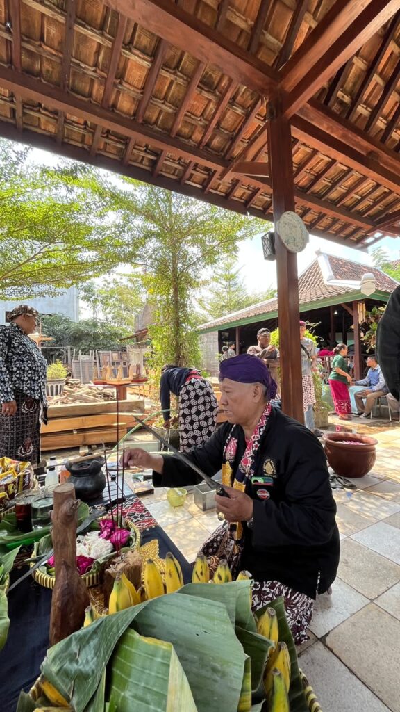 Elderly man in a purple headscarf prepares Balinese offerings with flowers and bananas under a wooden pavilion.