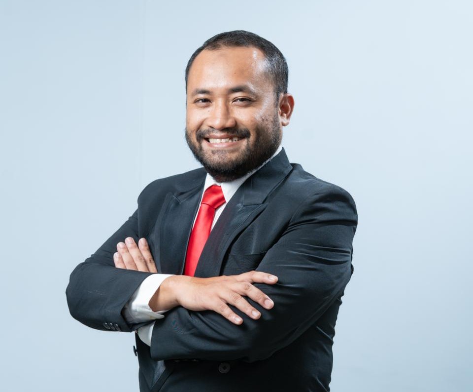Professional portrait of a smiling man in a dark suit and red tie with arms crossed against a light blue background.