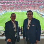 Two men in blazers pose for a photo in a stadium, with a full crowd and a green soccer pitch behind them in the stands
