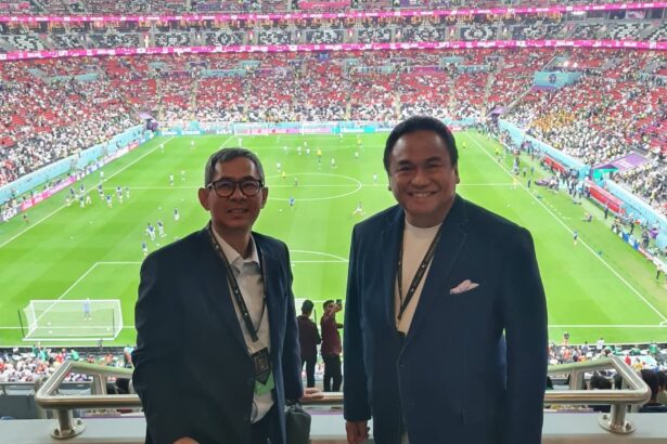 Two men in blazers pose for a photo in a stadium, with a full crowd and a green soccer pitch behind them in the stands