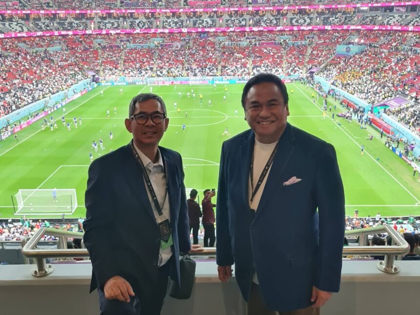 Two men in blazers pose for a photo in a stadium, with a full crowd and a green soccer pitch behind them in the stands