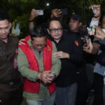 Man in a red vest being escorted in handcuffs by uniformed officers as reporters photograph the scene at night.