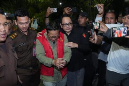 Man in a red vest being escorted in handcuffs by uniformed officers as reporters photograph the scene at night.