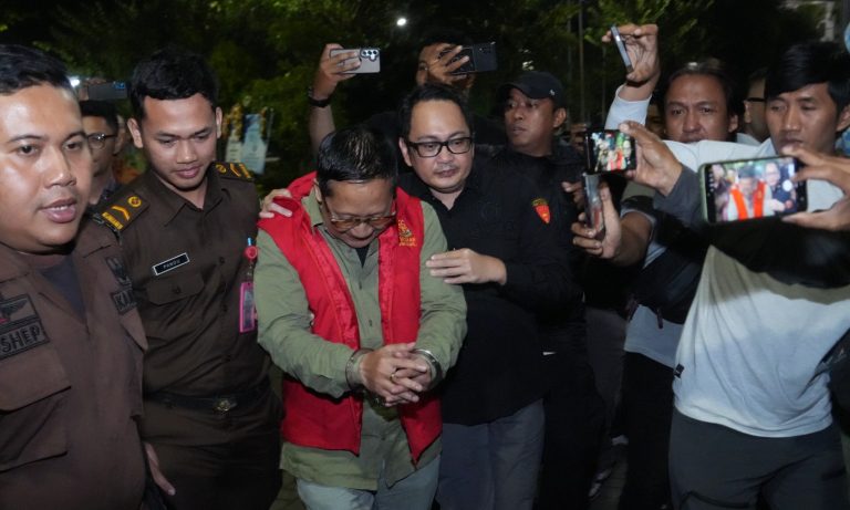 Man in a red vest being escorted in handcuffs by uniformed officers as reporters photograph the scene at night.