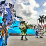 Performers in elaborate blue dragon costumes perform on a large outdoor stage with palm trees in the background.