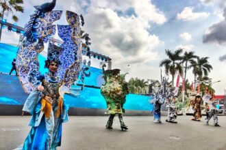 Performers in elaborate blue dragon costumes perform on a large outdoor stage with palm trees in the background.