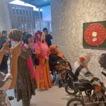 Group of people in a gallery admiring a framed circular rose artwork; some pose for photos, two women in wheelchairs nearby.