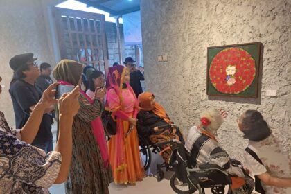 Group of people in a gallery admiring a framed circular rose artwork; some pose for photos, two women in wheelchairs nearby.