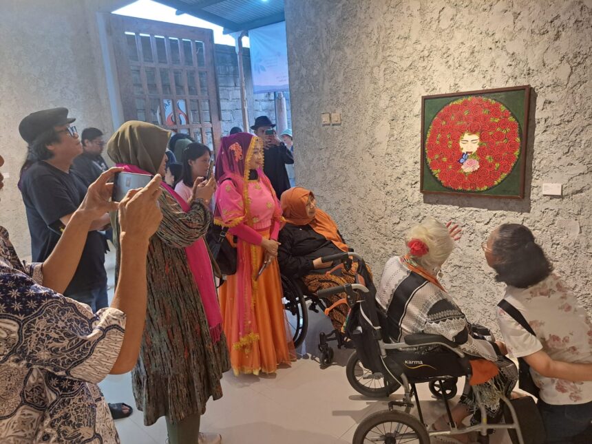 Group of people in a gallery admiring a framed circular rose artwork; some pose for photos, two women in wheelchairs nearby.