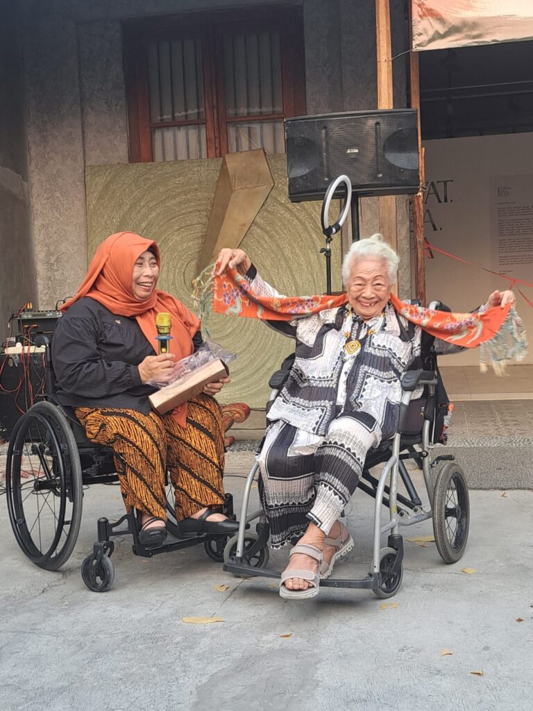 Two elderly women in wheelchairs share a joyful moment outdoors; one wearing an orange headscarf holds a microphone and a gift while the other lifts an orange patterned scarf, both smiling.