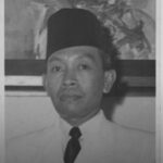 Black-and-white portrait of a man wearing a tall songkok hat, a white shirt, and a black tie, looking at the camera.