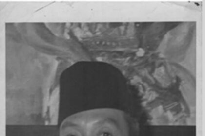 Black-and-white portrait of a man wearing a tall songkok hat, a white shirt, and a black tie, looking at the camera.
