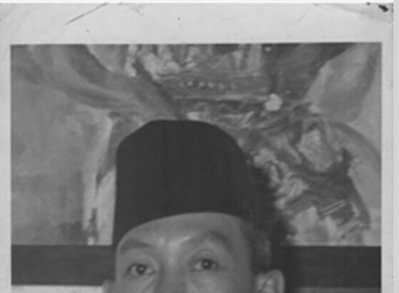 Black-and-white portrait of a man wearing a tall songkok hat, a white shirt, and a black tie, looking at the camera.