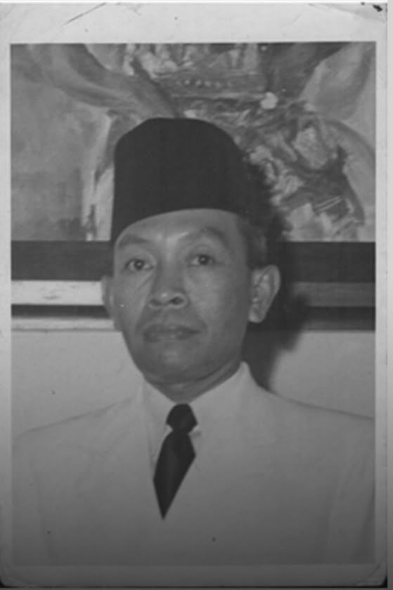 Black-and-white portrait of a man wearing a tall songkok hat, a white shirt, and a black tie, looking at the camera.